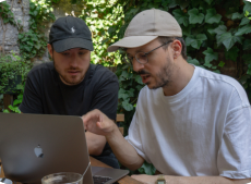 2 employees in white and black T-shirts pointing at the MacBook: Two of the Once Advertisements employees, Krisztian Mezei and Kristof Menesi sitting in Lumen Café discussing business matters.